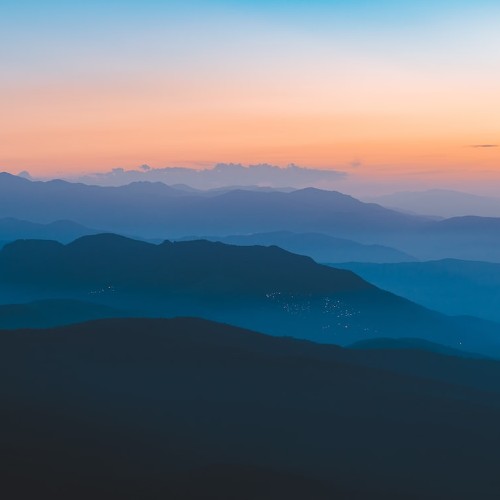 A hazy sunset across fog shrouded, waving hills, a faint trace of civilization glimmers among them.