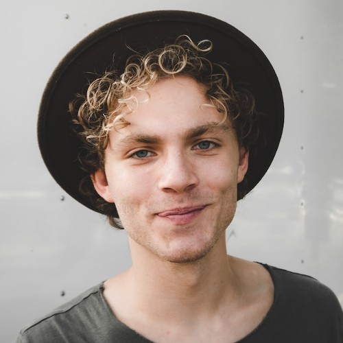 Portrait of a man with curly, dirty blond hair peeking out from up a mid-brimmed hat.  He wears a playful smirk.